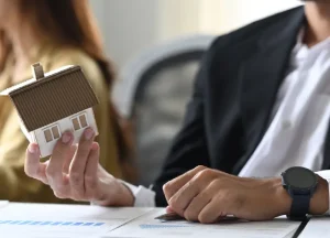 Businessperson holding a small house model while reviewing real estate investment charts on a laptop.