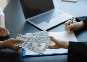 Fix and Flip Loans vs. Hard Money Loans: Which One Fits Your Strategy? 3 Close-up of a stack of US hundred-dollar bills being passed from one person's hands to another over a desk, with a laptop and a contract being signed in the background.