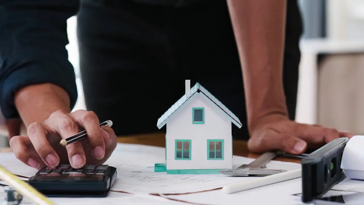 Investor calculating fix and flip profit margins using a calculator, blueprints, and renovation measurements at a project planning table.