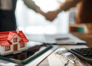 DSCR Loans for First-Time Investors: What You Need to Get Started 3 A close-up view of a model house with a red roof on a desk with a calculator and eyeglasses, with a blurred handshake between two people in the background, signifying a real estate deal.